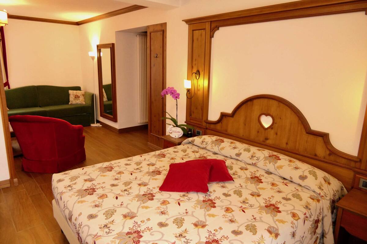 A hotel room with a bed, a green sofa, a red armchair, and a wooden headboard with a heart-shaped design.