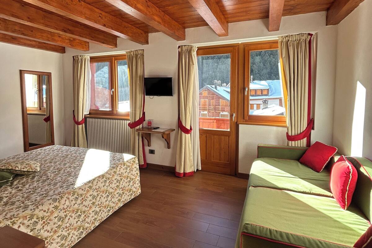 A room with wooden beams on the ceiling, a bed with floral bedding, a sofa with red cushions, and a window with curtains.