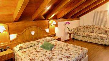 Ein Schlafzimmer mit Holzbalkendecke, einem blumigen Bettbezug, einem grünen Kissen, einem Nachttisch, einer Lampe, einem blumengemusterten Sofa und einem kleinen Tisch mit Blumenvase.