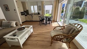 Un salon moderne avec un canapé, une table basse et un fauteuil en osier. On y trouve également un coin repas avec une table et des chaises, et une cuisine à l’arrière-plan.