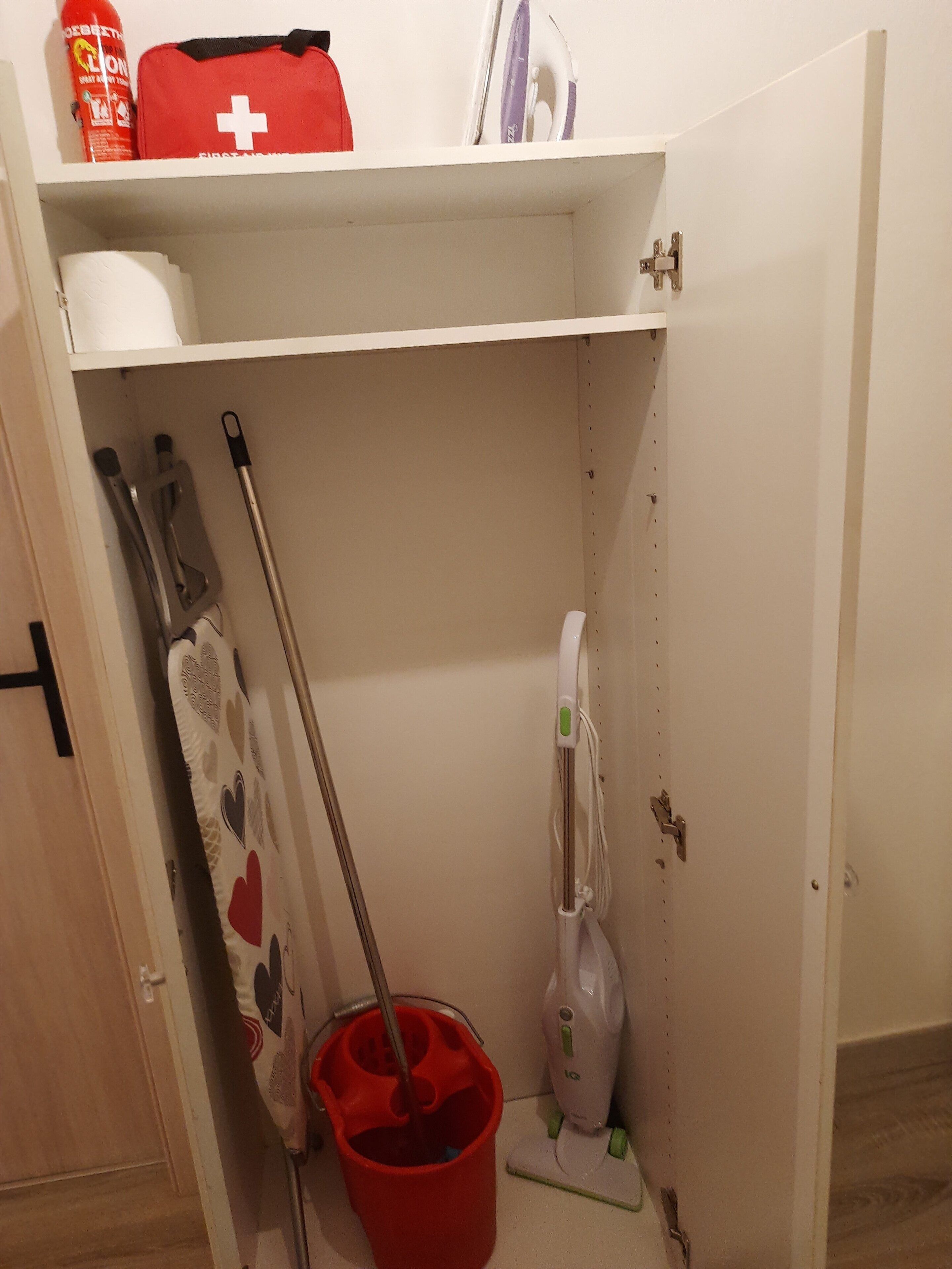 A small storage space with a red first aid kit, a mop, a bucket, and a vacuum cleaner.