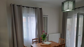 A dining area with a wooden table and chairs, a vase with flowers, and a view of the outside through large windows with sheer curtains.