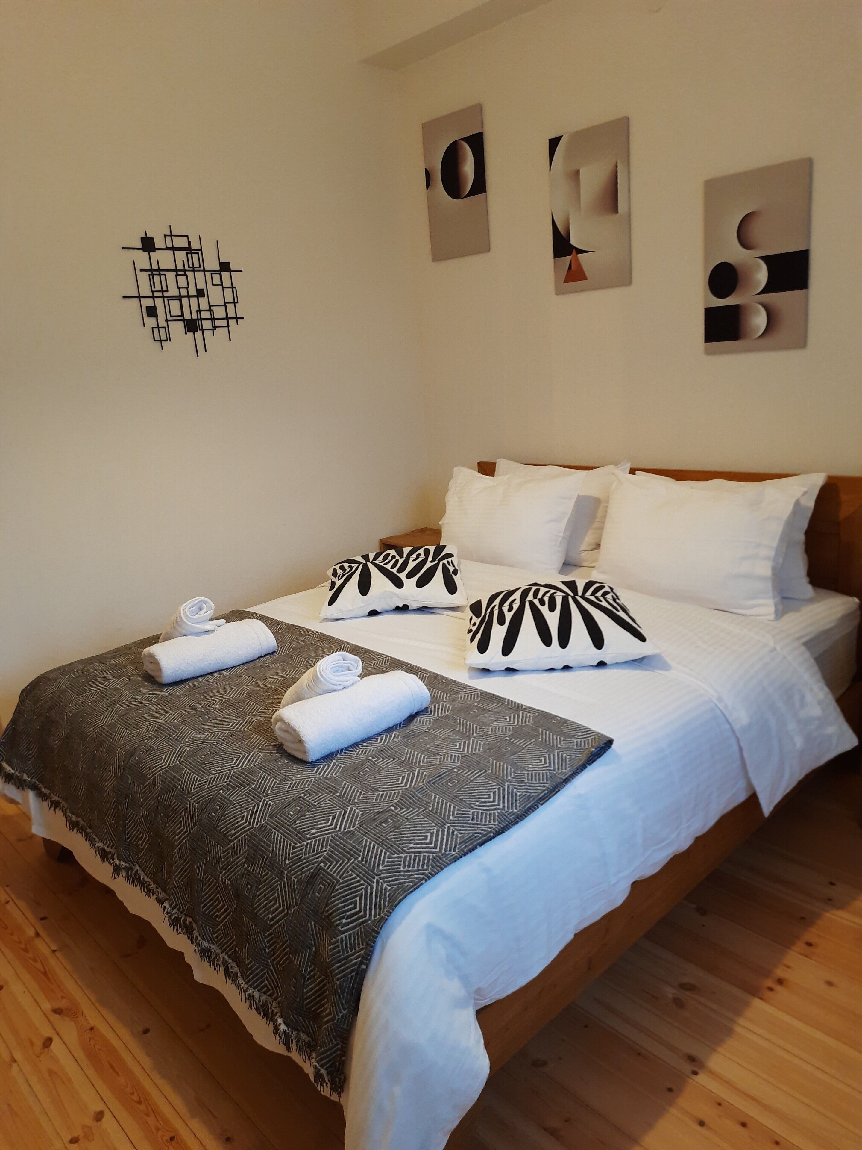A neatly made bed with white linens and patterned pillows, a dark patterned throw blanket, and two rolled towels on the foot of the bed.