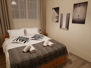 A neatly made bed with patterned pillows and a geometric throw blanket, a bedside table with a lamp, and abstract wall art. - AkisPlace. 
3 bedroom apt fully rennovated, cozy and comfortable place to stay. (Ioannina)