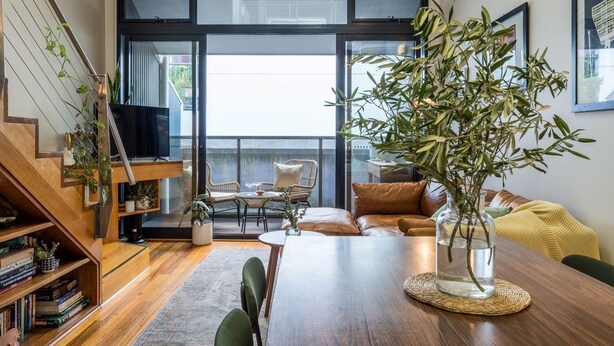 A modern living room with a wooden staircase, a large window, and a dining area with a table and chairs. - StayCentral - Brunswick Loft - Michael Street, Brunswick - 1 Bedroom, 1 Bed, 1.5 bathroom, 1 Parking, Pet Friendly (Brunswick)