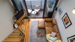 A modern living room with a leather sofa, a coffee table, and a TV. There is a balcony with outdoor seating and plants. - StayCentral - Brunswick Loft - Michael Street, Brunswick - 1 Bedroom, 1 Bed, 1.5 bathroom, 1 Parking, Pet Friendly (Brunswick)