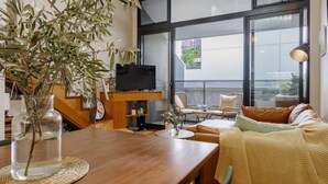 A modern living room with a wooden table, a sofa, and a TV. There is a balcony with wicker chairs and a view of the outside. - StayCentral - Brunswick Loft - Michael Street, Brunswick - 1 Bedroom, 1 Bed, 1.5 bathroom, 1 Parking, Pet Friendly (Brunswick)