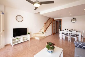 A living room with a flat-screen TV, a staircase, a dining area with a table and chairs, and a ceiling fan.