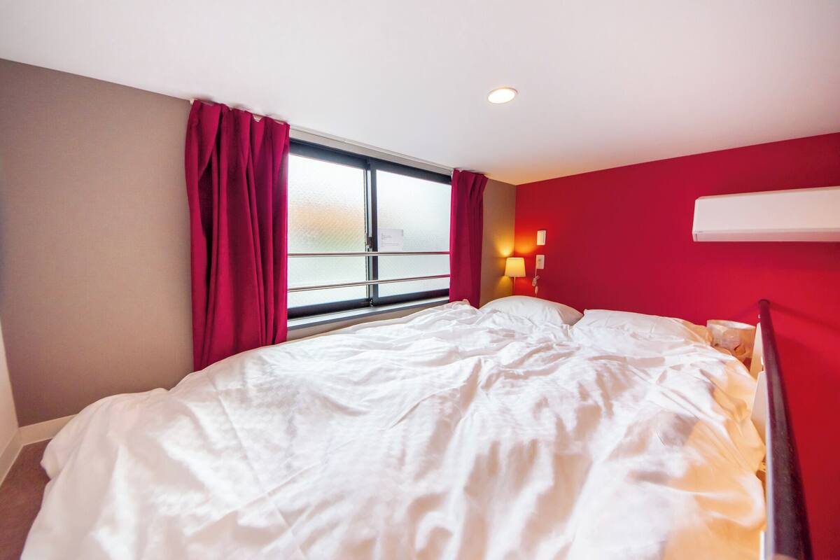 A bedroom with a large bed, red curtains, a window, and a wall-mounted air conditioner.