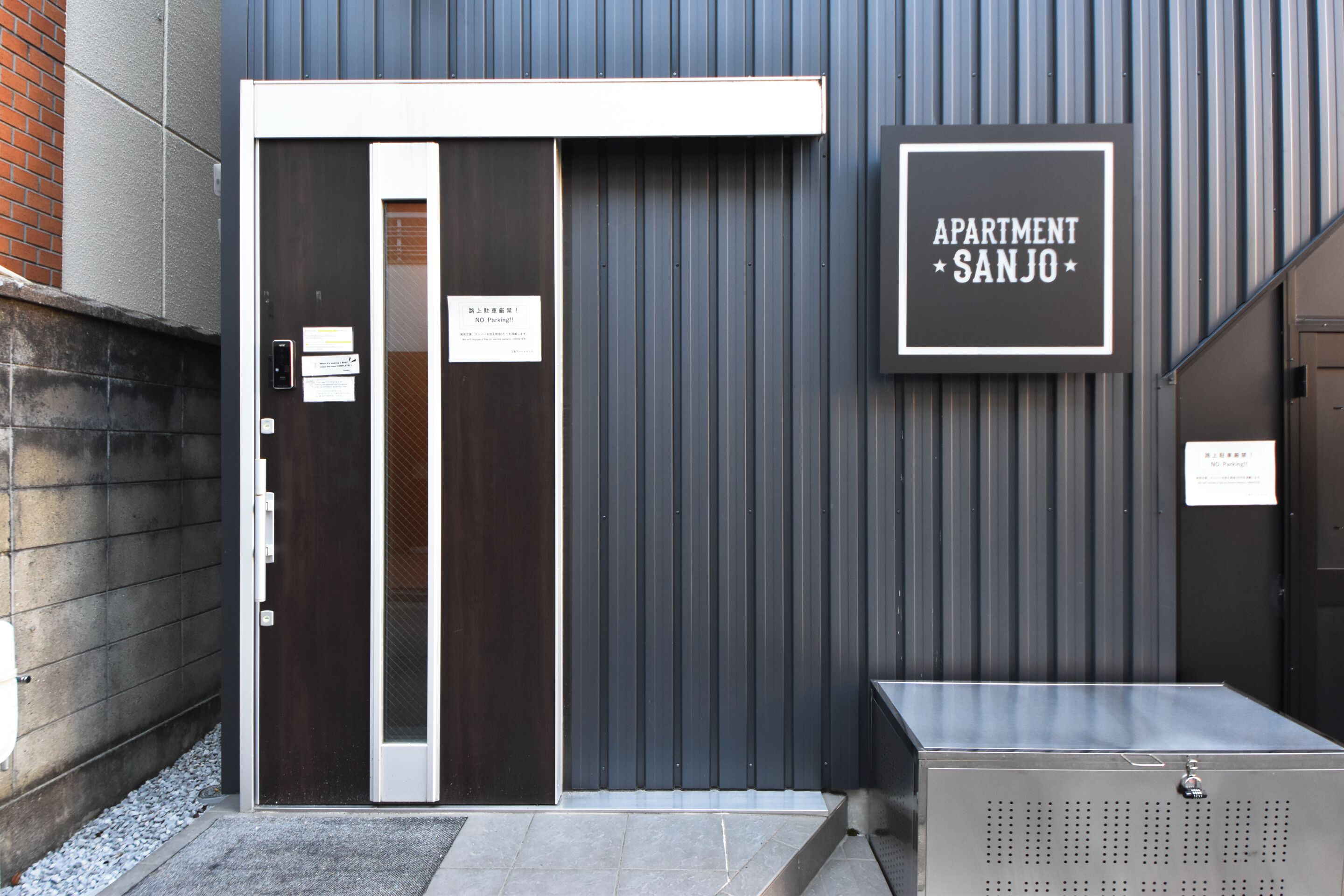 A modern building entrance with a metal door, a sign reading "APARTMENT SANJO," and a small metal box on the ground.