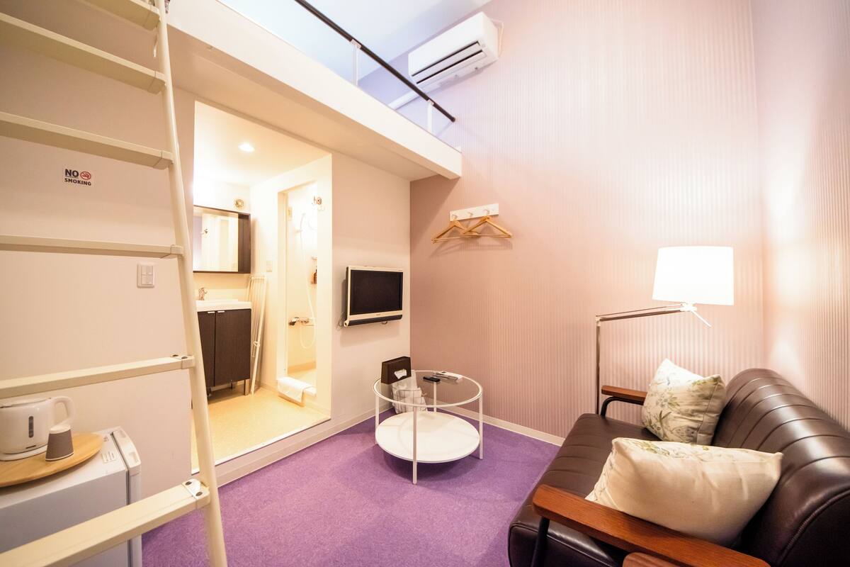 A hotel room with a purple carpet, a brown leather sofa, a round glass table, a TV, a hanging clothes rack, and a white wall with a 'No Smoking' sign.