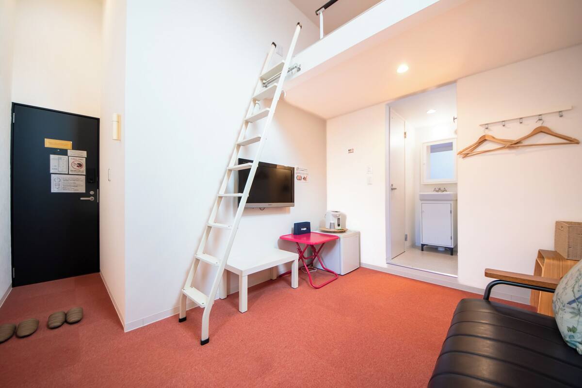 A compact room with a white desk, a small table with a red chair, a TV mounted on the wall, a black door, and a red carpeted floor.