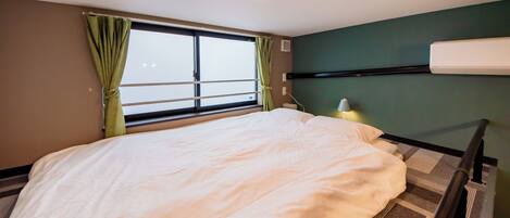 Habitación de hotel con cama, ventana con cortinas, mesita de noche con lámpara y aire acondicionado instalado en la pared.