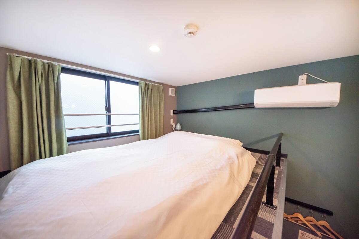 A hotel room with a bed, a window with curtains, an air conditioning unit, and a wall-mounted shelf.