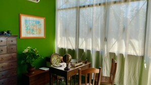 A room with a green wall, a wooden dresser, a desk with a chair, a potted plant, and a window with sheer curtains.