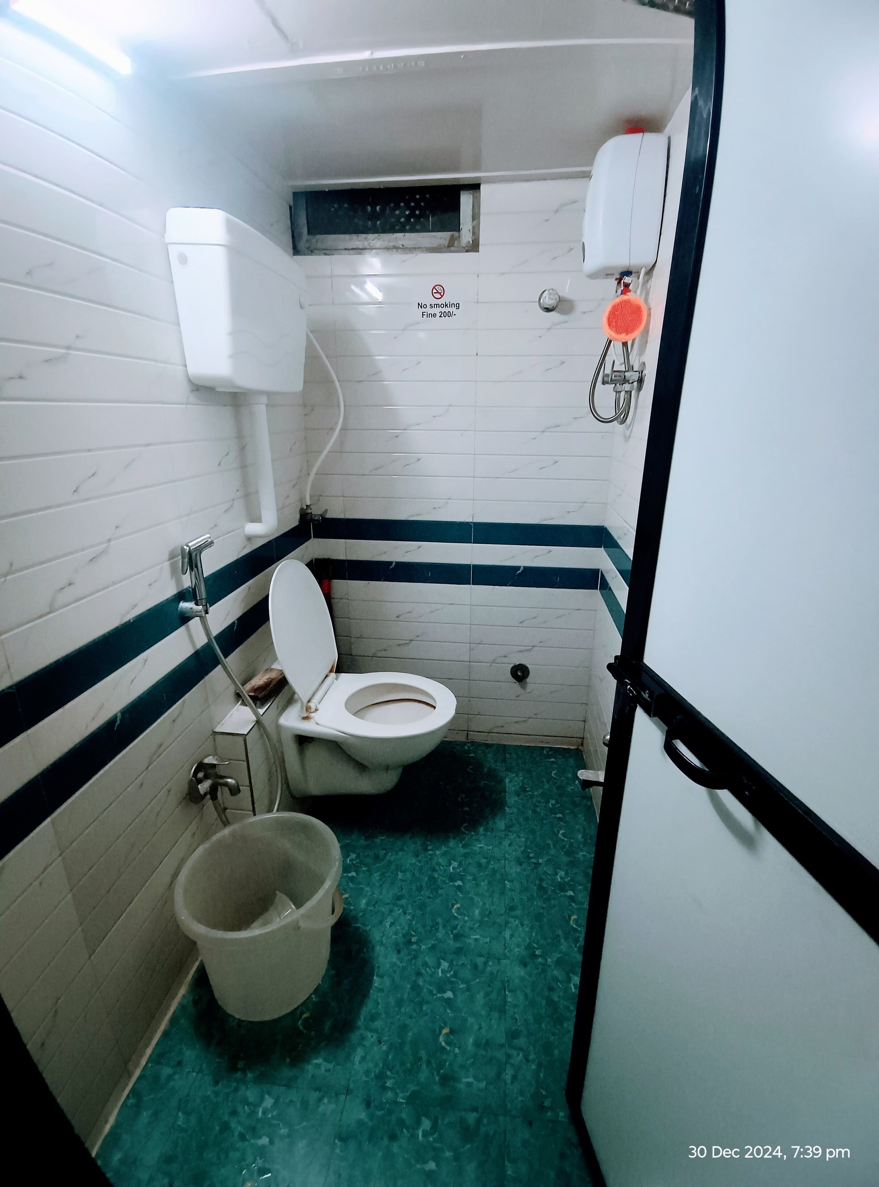 A small, narrow bathroom with a toilet, a bucket, and a shower area.