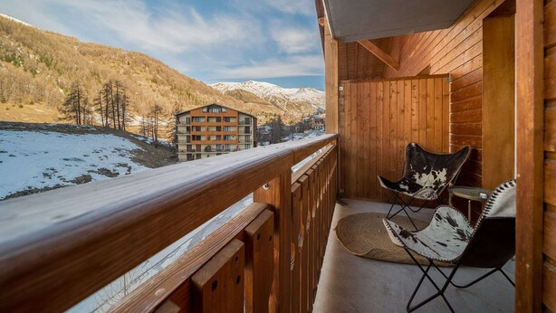 A wooden balcony with a view of a mountain and a building. There is a chair and a small table on the balcony. - Studio in France with Mountain Views & Pool (Allos)