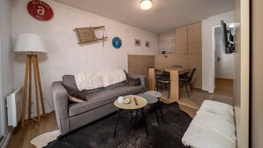 A compact living space with a grey sofa, a small round table, a floor lamp, a dining area with a chair, and a TV mounted on the wall. - Studio in France with Mountain Views & Pool (Allos)