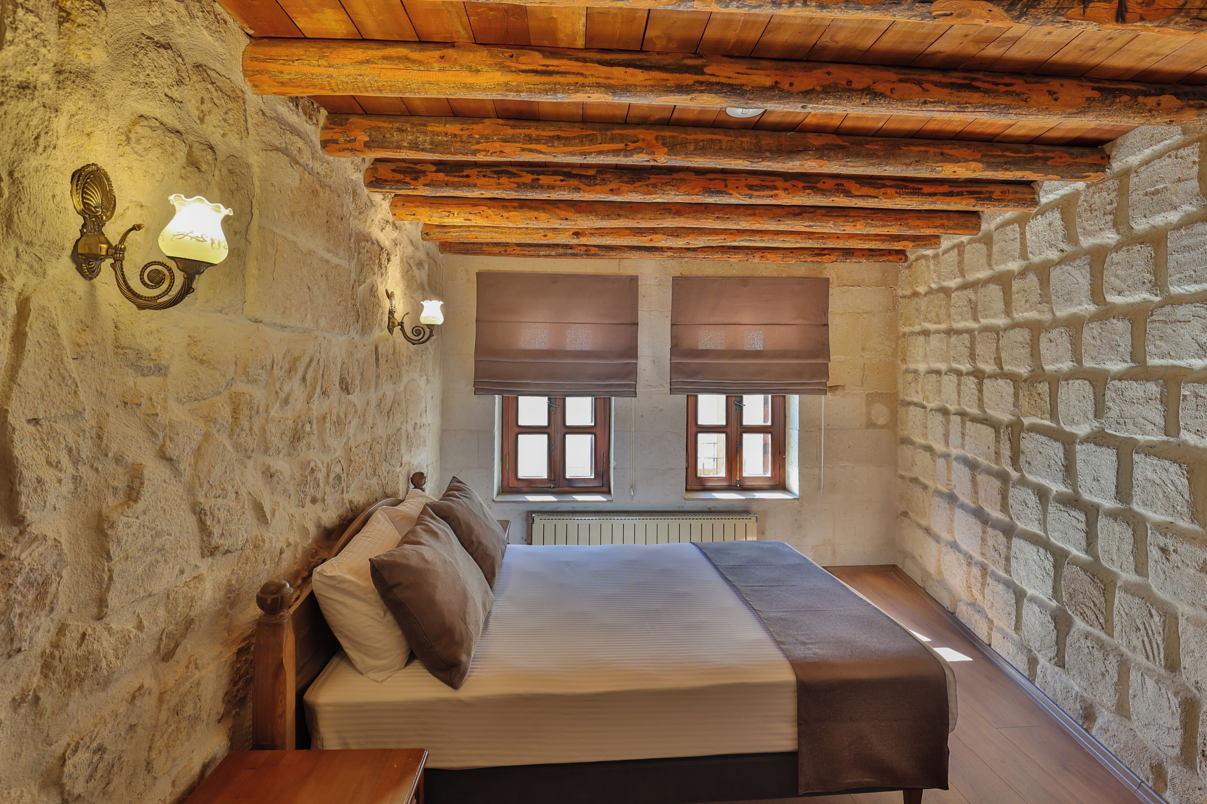 Une chambre avec un mur en pierre, des poutres en bois, un lit avec des oreillers et deux fenêtres équipées de stores.