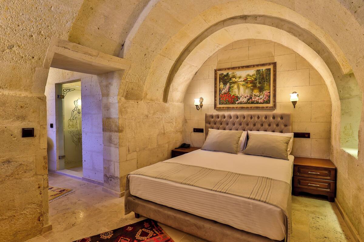 Quarto com cama grande, mesa de cabeceira de madeira, abajur na parede e quadro sobre a cama.