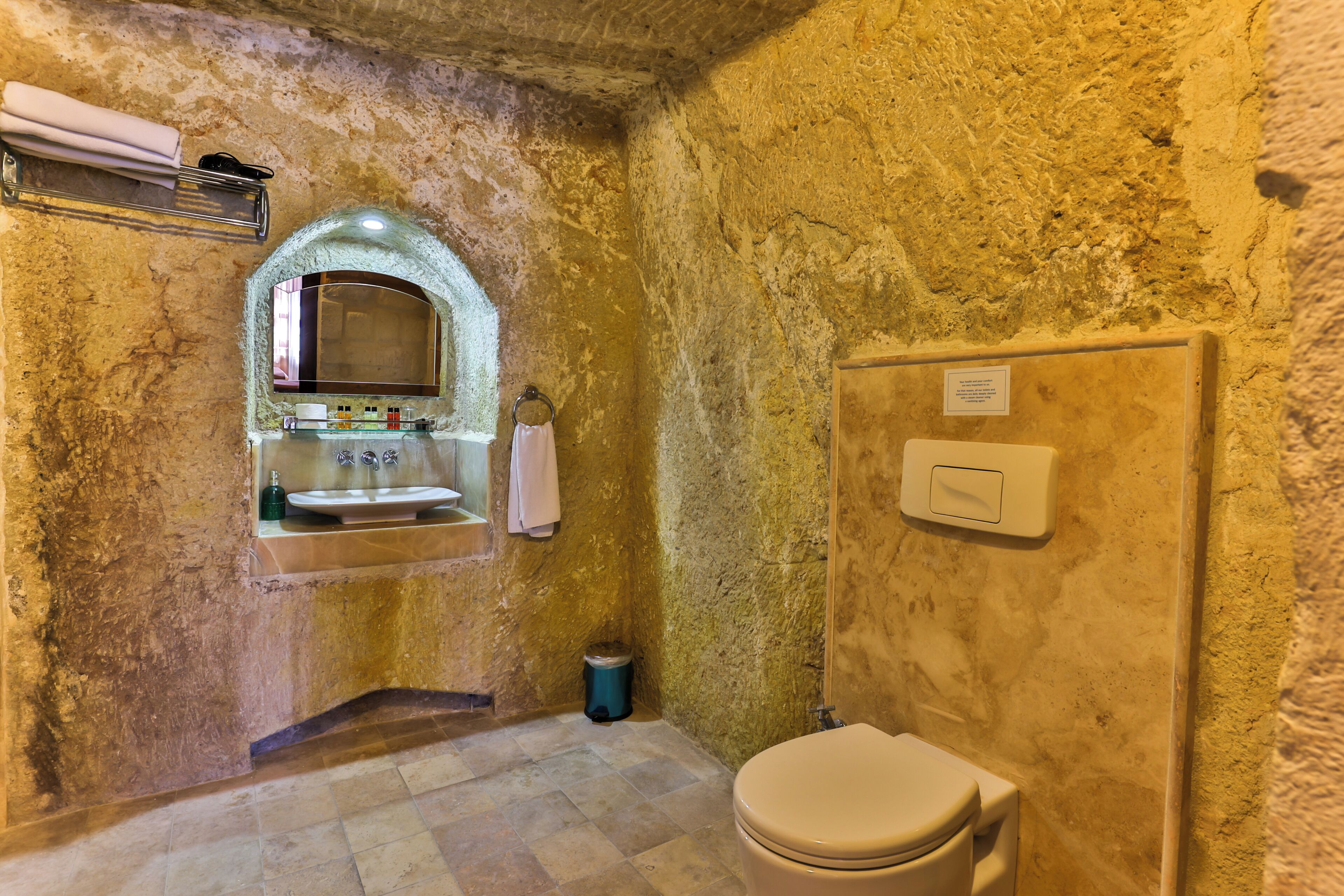 A bathroom with a stone wall, a sink with a mirror, a toilet, and a trash bin.