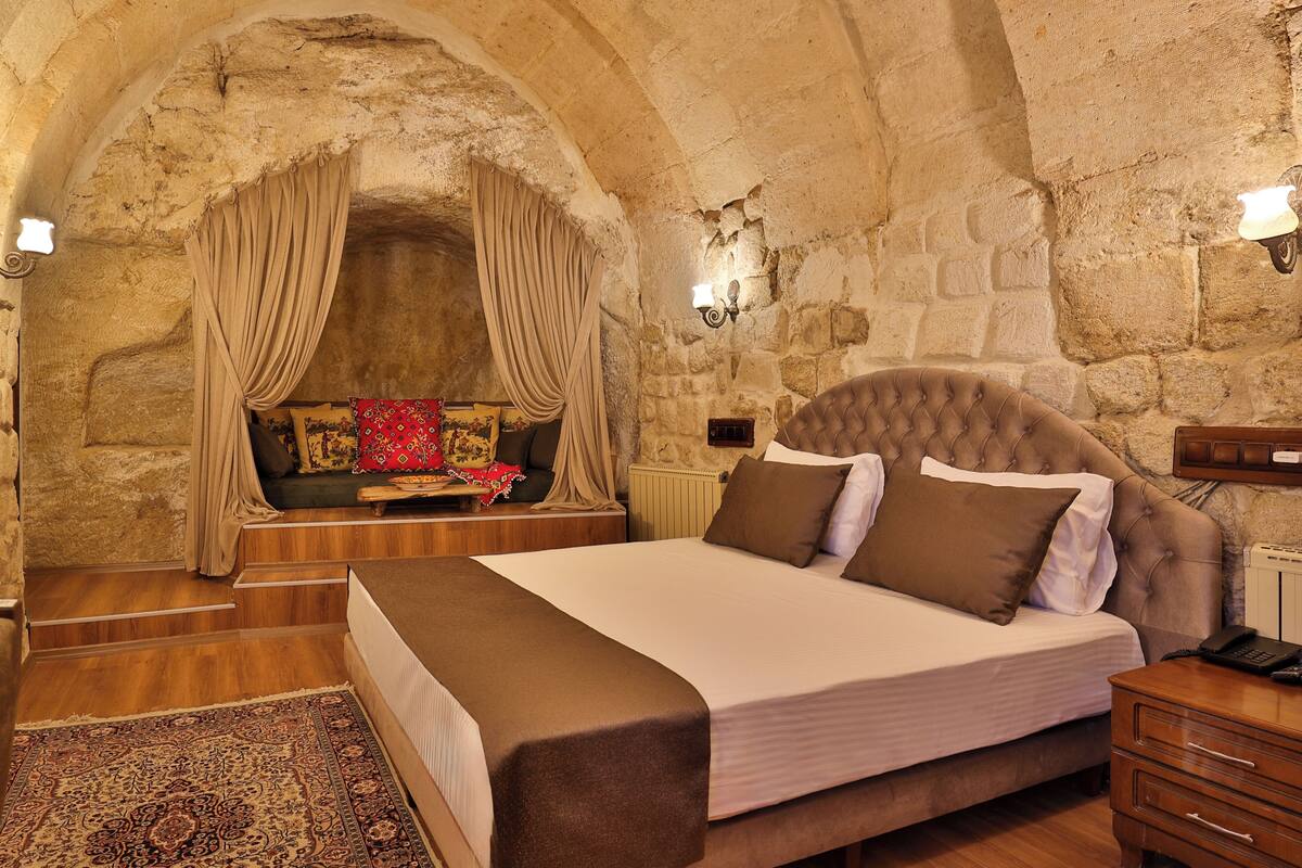 Quarto com parede de pedra, cama com cabeceira marrom, mesa de cabeceira de madeira e travesseiro decorativo vermelho.