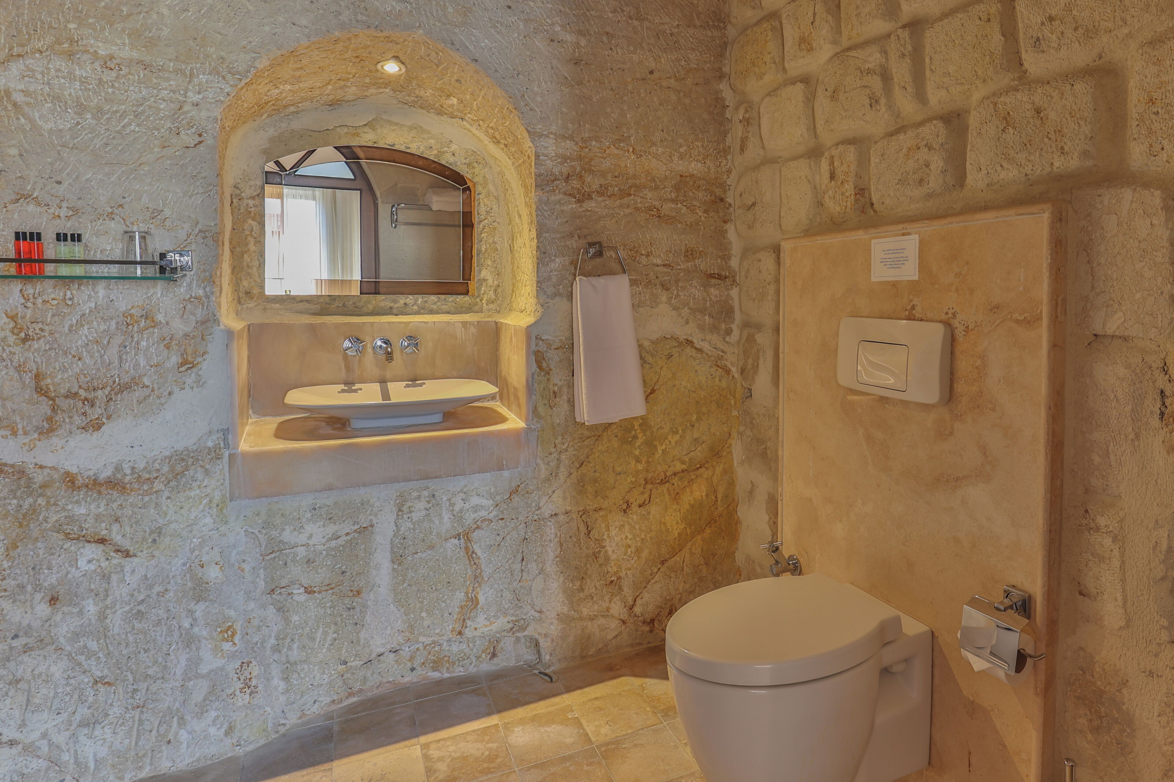 A bathroom with a stone wall, a sink with a mirror, a toilet, and a towel rack.