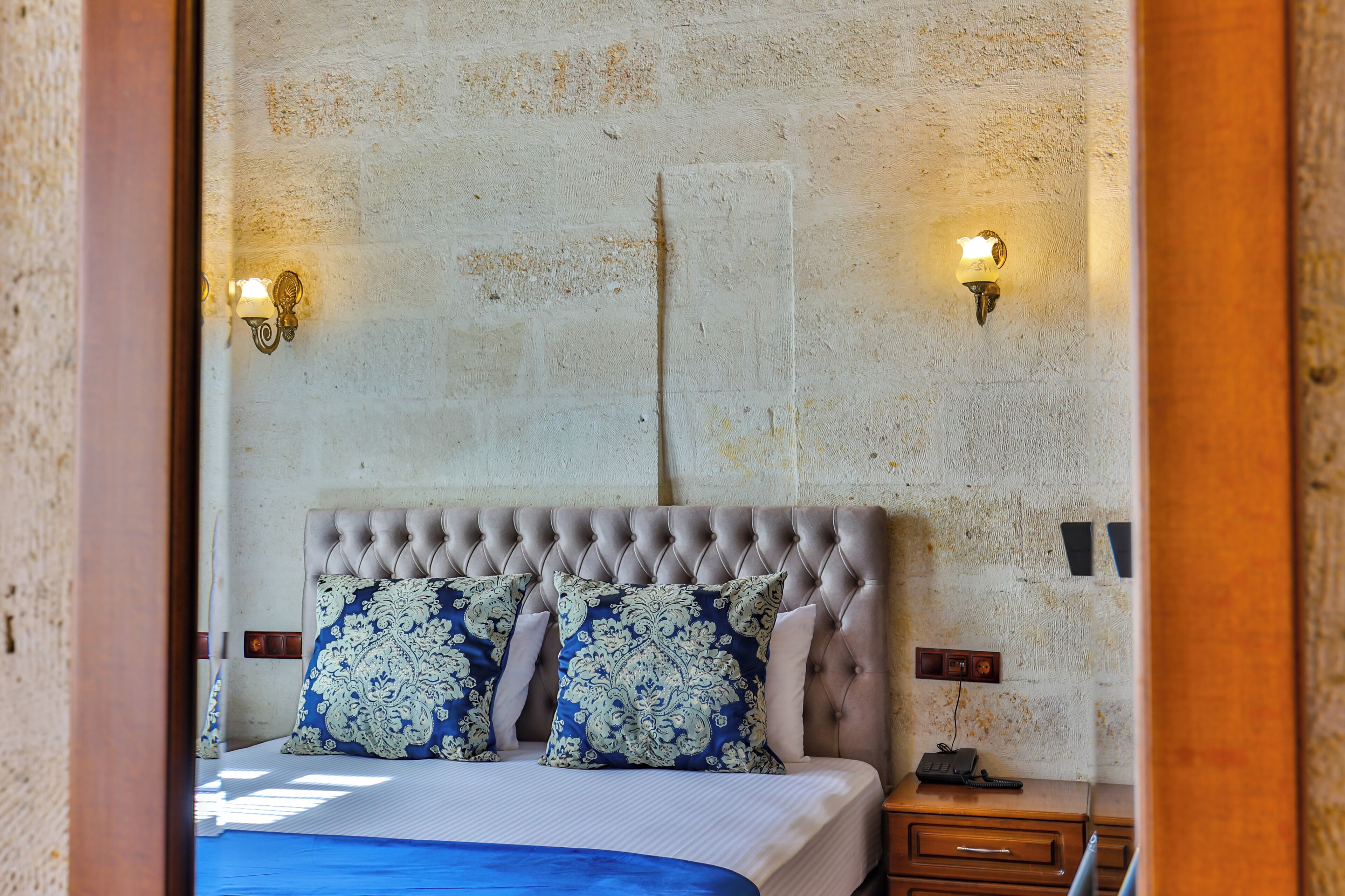 A bedroom with a tufted headboard, a bed with blue and white patterned bedding, two wall-mounted light fixtures, and a nightstand with a telephone.