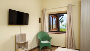 Una stanza con un letto, una televisione a muro, una finestra con tende, una poltrona verde e una piccola mensola con libri.