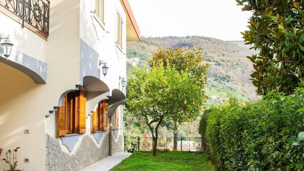 A building with a stone facade, wooden shutters, and a paved pathway leading to a lush green garden with a tree and a hedge. - 🌺Villa dei Fiori – Elegance, Nature & Serenity on the Sorrento Coast (Vico Equense)