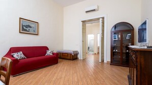 Un salón con sofá rojo, suelo de madera, un mueble con televisor y una puerta que da a otra habitación.