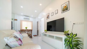 A modern living room with a sofa, a TV mounted on a white entertainment unit, and a dining area with a table and chairs. - Beachfront 2BR Pool Villa DelMar Kamala (Kamala)