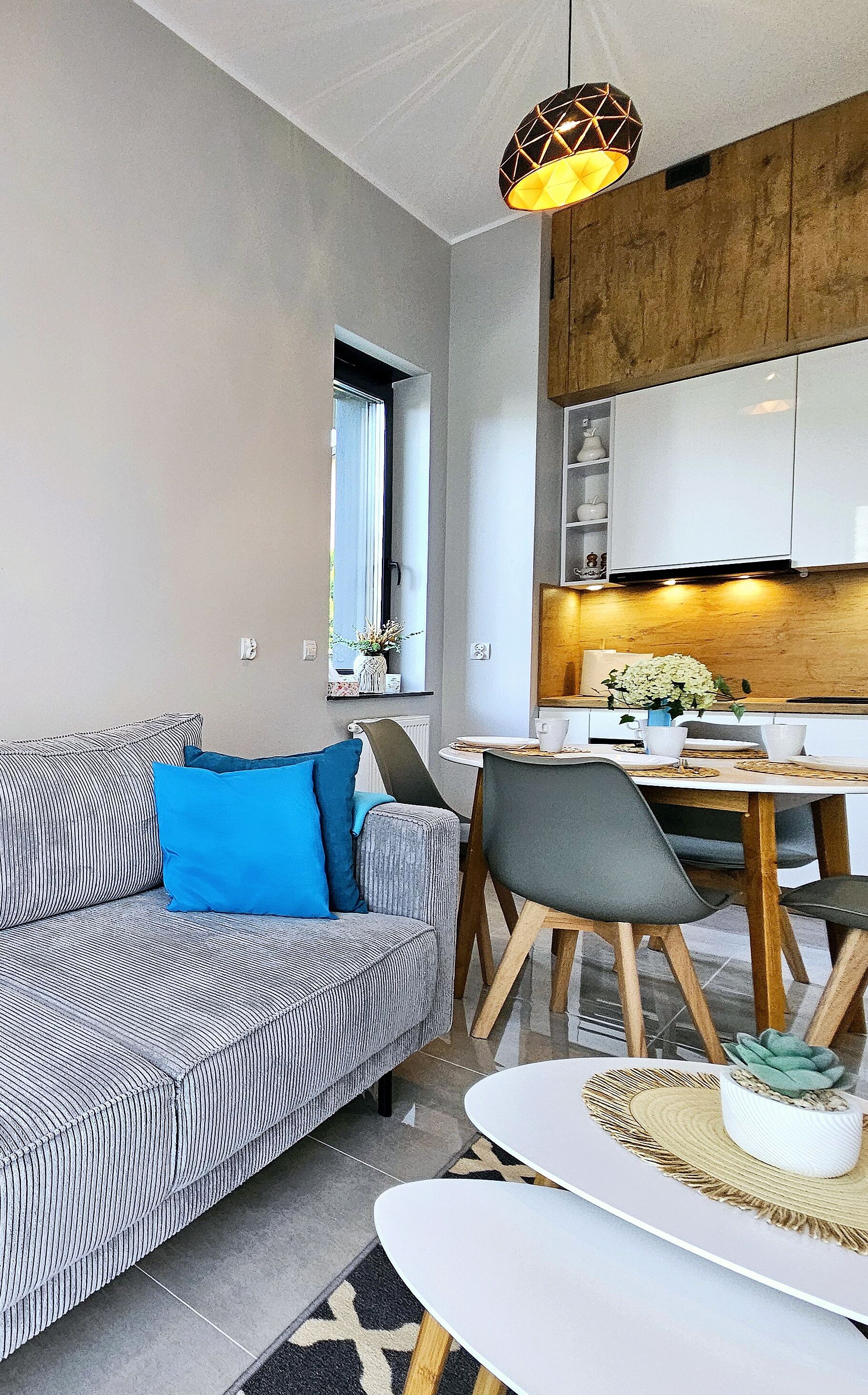 A modern living room with a sofa, a dining table, and chairs. There is a kitchen area with cabinets and a window.
