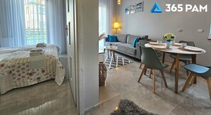 A modern apartment with a bed, sofa, dining table, and chairs. There are curtains, a lamp, and a small plant.