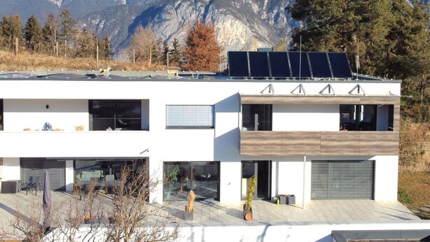 Ein modernes, zweistöckiges Gebäude mit Solaranlage auf dem Dach, großen Fenstern und Blick auf schneebedeckte Berge.