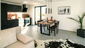 Eine moderne Küche mit dunklen Schränken, eine Essecke mit einem Holztisch und ein Wohnzimmer mit einer Couch und einem Kuhfellteppich.