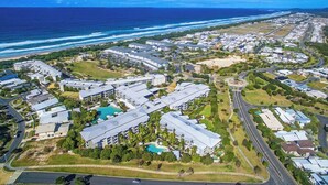 An aerial view of a coastal residential area with multiple buildings, roads, and a beach. - The Sand Resort Kingscliff - One Bed Pool View (3208) (Tweed)