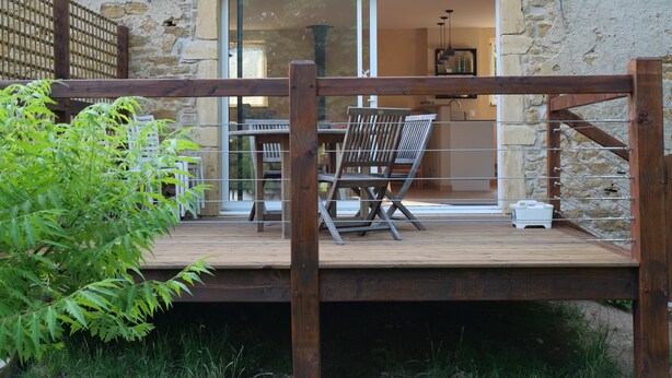A wooden deck with a table and chairs, a glass door leading to a kitchen, and a stone wall. - Le Clos de la Ferdière - charming gîte near Cluny (Navour-sur-Grosne)