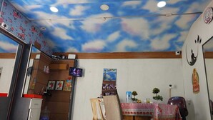 A room with a ceiling painted to resemble clouds, a small table with a floral tablecloth, a chair, and a television mounted on the wall. - Yogi Golden Resort (Mysore)