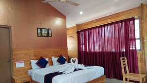 A wooden-framed bed with white bedding and decorative pillows, a ceiling fan, and a window with red curtains. - Yogi Golden Resort (Mysore)