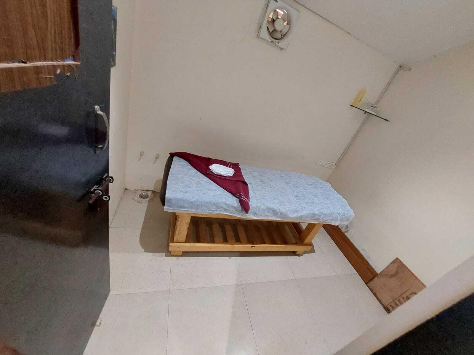 A small, single-bed room with a wooden bed frame, a white mattress, and a red blanket. There is a small wooden stool beside the bed and a white wall with a small shelf.