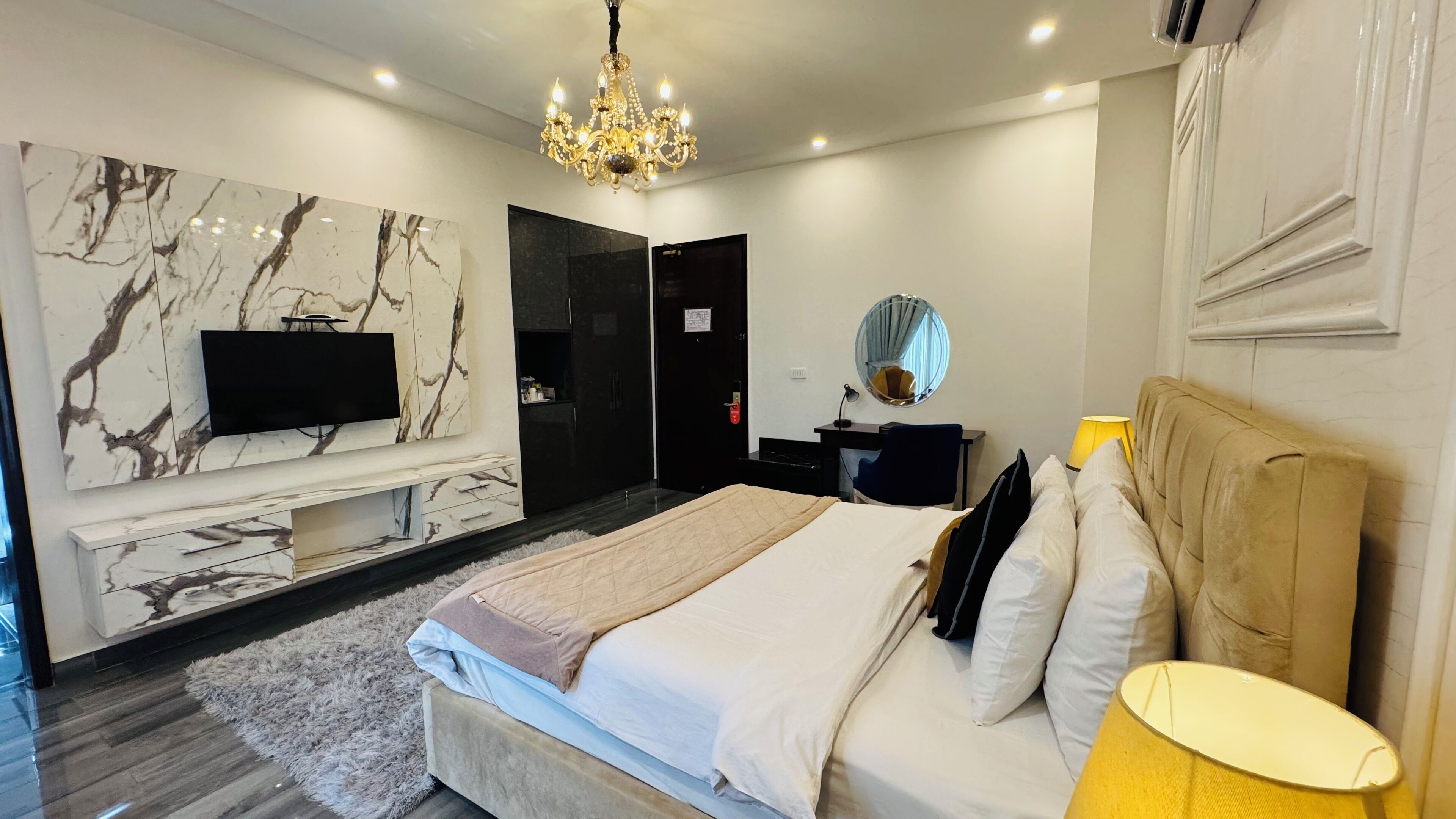 A modern bedroom with a bed, a TV mounted on a marble wall, a chandelier, a mirror, and a bedside table.