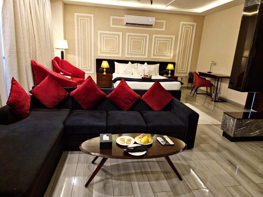 A hotel room with a bed, a sofa with red cushions, a coffee table with breakfast items, and a TV.