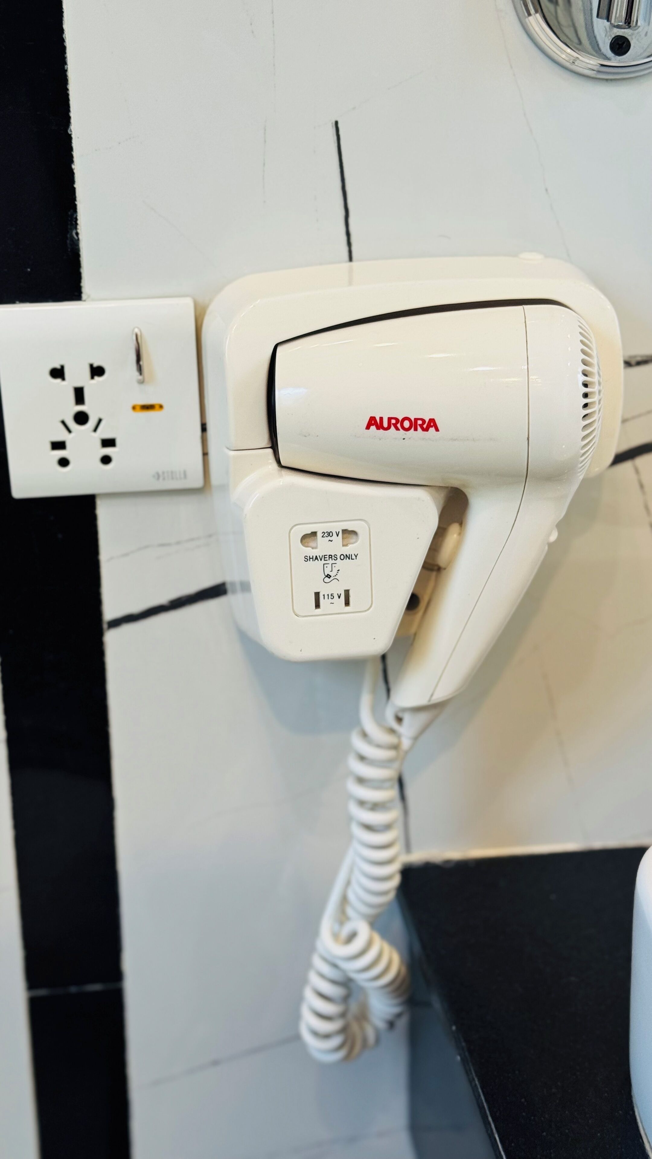 Hiustenkuivaaja, jossa on pistoke, joka on kytketty pistorasiaan. Laitteessa on merkinnät »AURORA» ja »VAIN PARRANAAJINTEN KÄYTTÖÖN».