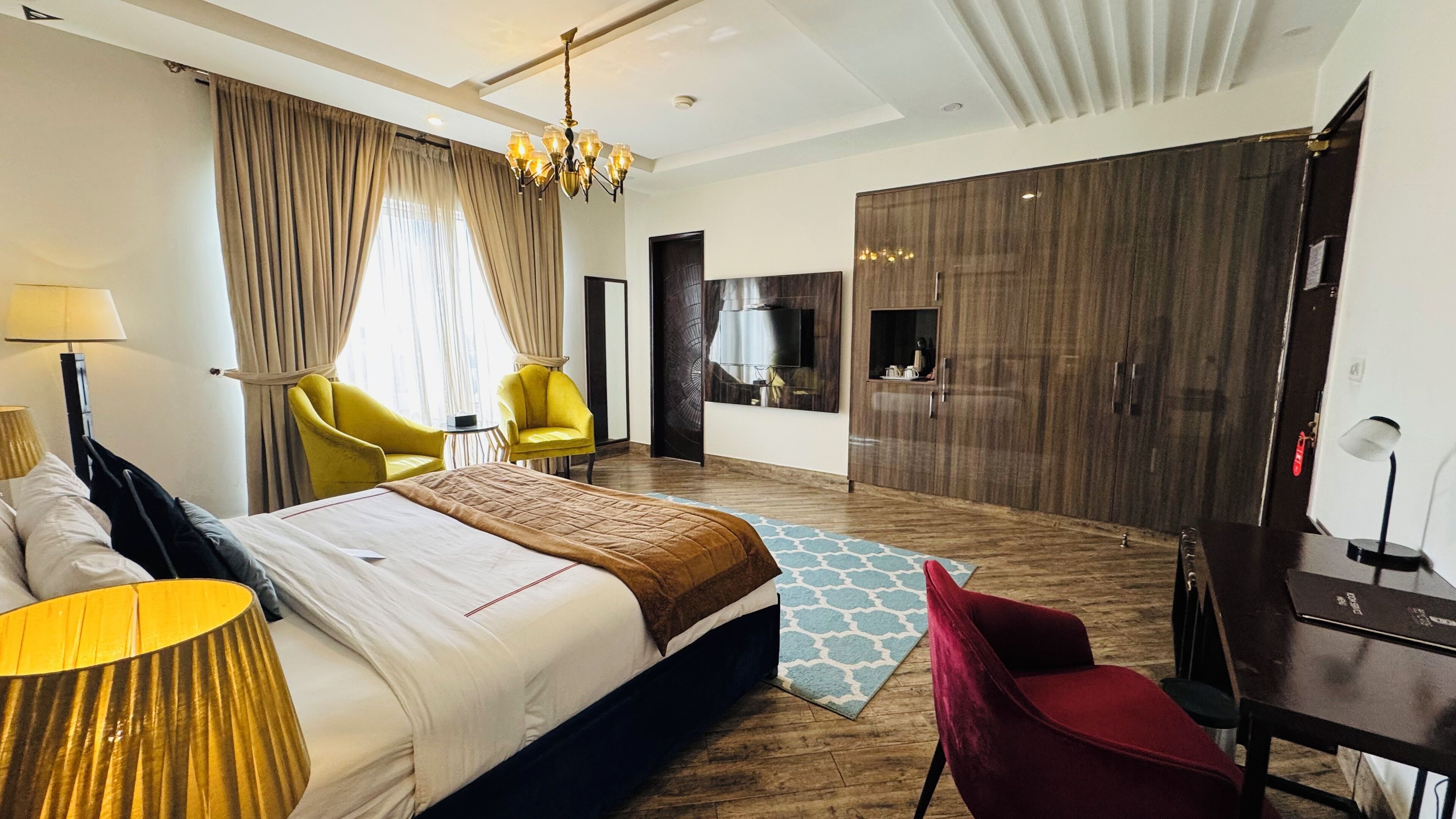 Une chambre d’hôtel avec un grand lit, deux fauteuils jaunes, une chaise rouge, un plancher en bois, un lustre, une télévision et un mini-bar.
