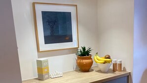 Un piano di lavoro in cucina con un'opera d'arte incorniciata, una pianta in vaso, una ciotola di frutta e una pila di scatole da tè.