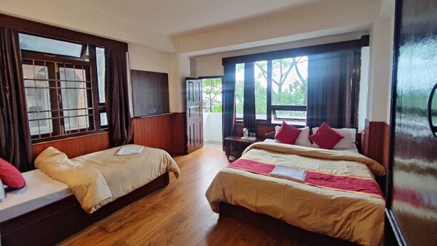 A hotel room with two single beds and one double bed, wooden flooring, large windows with curtains, and a view of greenery outside.