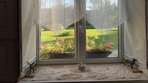 A room with a large window featuring a view of a garden with flowers and a candle on the windowsill.
