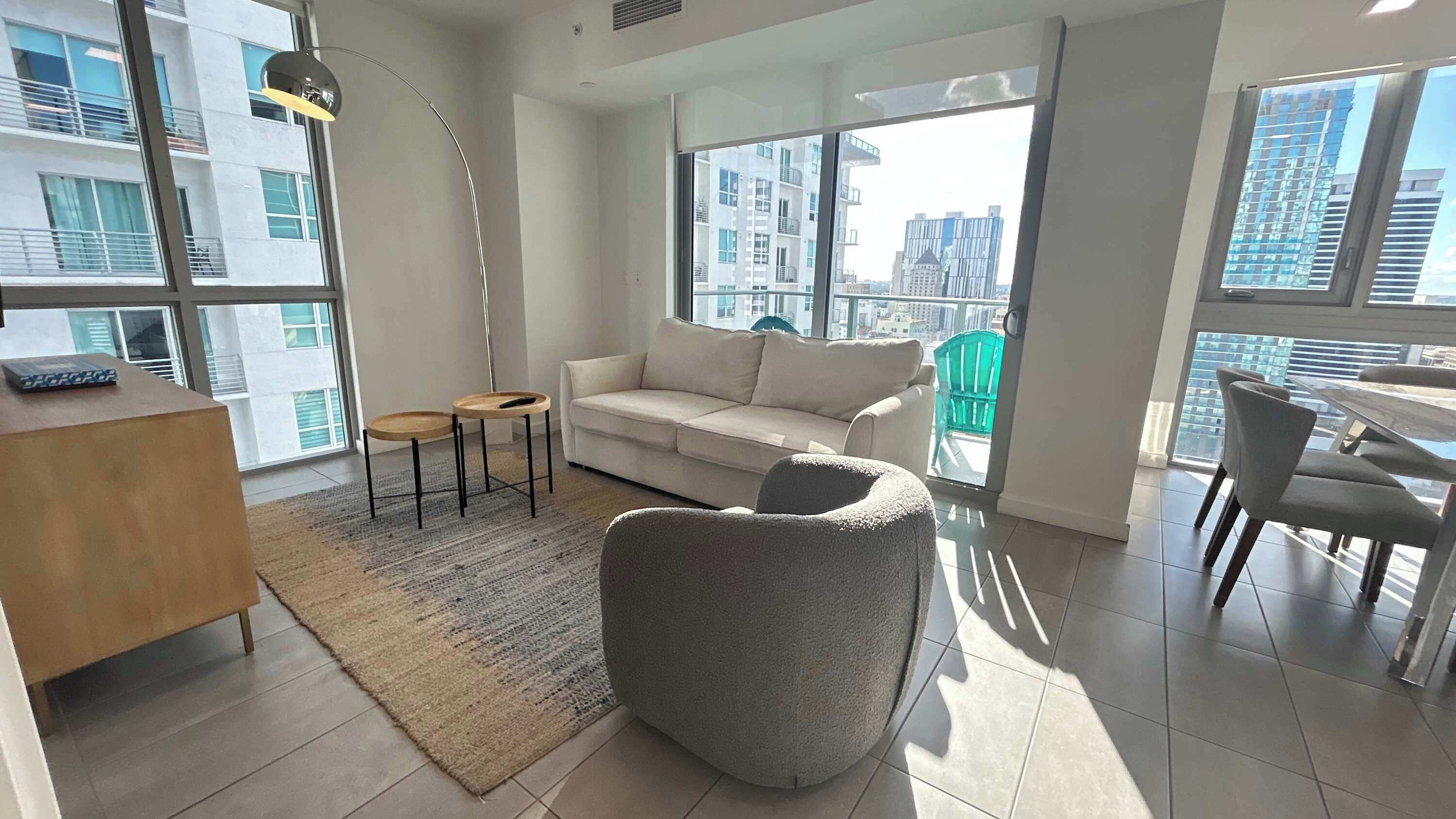 A modern living room with a sofa, a round table, and a chair. There is a large window with a view of buildings.