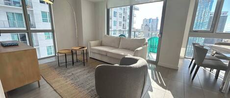 A modern living room with a sofa, a round table, and a chair. There is a large window with a view of buildings.